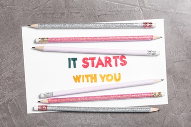 Sheet of paper with phrase It Starts With You and pencils on grey table, top view