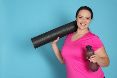 Happy overweight woman with bottle of water and yoga mat on light blue background