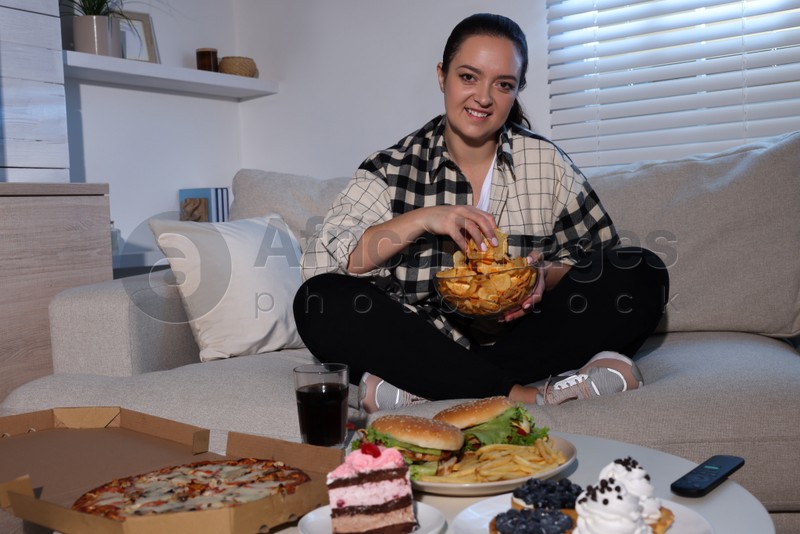 Happy overweight woman with chips on sofa at home. Unhealthy food