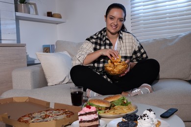 Photo of Happy overweight woman with chips on sofa at home. Unhealthy food