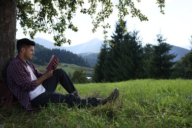Handsome man reading book under tree on meadow in mountains