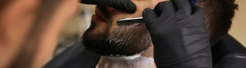 Professional hairdresser shaving client with straight razor in barbershop, closeup. Banner design