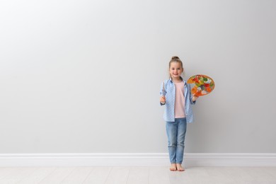 Little girl with brush and paint palette near light grey wall indoors. Space for text