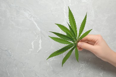Woman holding fresh green hemp leaf at marble table, closeup. Space for text