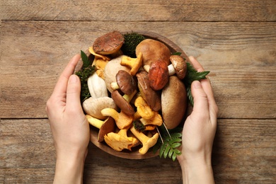 Photo of Top view of woman holding bowl with different wild mushrooms on wooden background, closeup