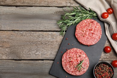 Raw hamburger patties with rosemary, tomatoes and peppercorns on wooden table, flat lay. Space for text