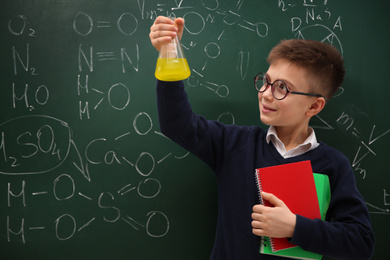 Schoolboy holding flask and notebooks near chalkboard with chemical formulas