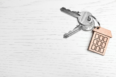 Keys with trinket in shape of house on white wooden background, top view and space for text. Real estate agent services