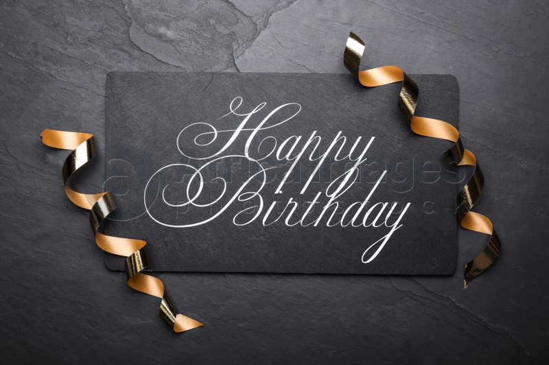 Slate board with greeting HAPPY BIRTHDAY and serpentine streamers on black background, top view