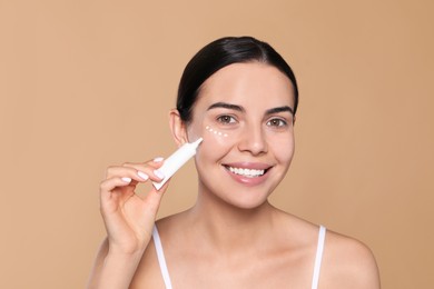 Beautiful young woman applying cream on skin under eye against beige background