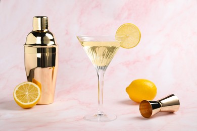 Photo of Martini glass of refreshing cocktail with lemon slice and fresh fruits near shaker on pink marble background