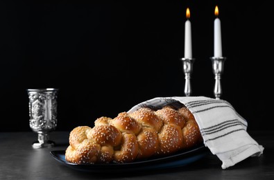 Homemade braided bread with sesame seeds, goblet and candles on dark grey table. Traditional Shabbat challah