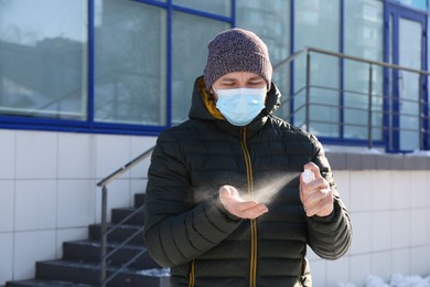 Photo of Man in protective mask spraying antiseptic onto hand outdoors