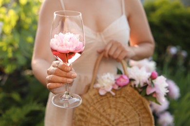 Woman with glass of rose wine and straw bag in peony garden, closeup