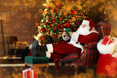 Santa Claus reading book near decorated Christmas tree indoors