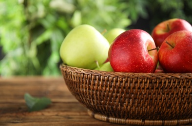 Photo of Different ripe apples in wicker bowl on wooden table against blurred background, closeup