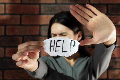 Photo of Domestic violence concept. Unhappy woman holding paper with written word Help near brick wall, focus on hands