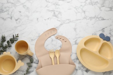 Flat lay composition with silicone baby bib and plastic dishware on white marble background. Space for text