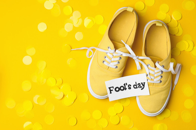 Shoes tied together, note with phrase FOOL'S DAY and confetti on yellow background, flat lay. Space for text