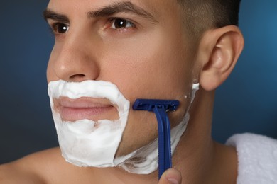 Handsome man shaving with razor on blue background, closeup