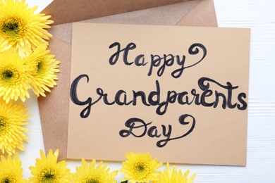 Beautiful yellow flowers, envelope and card with phrase Happy Grandparents Day on white wooden table, flat lay