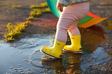 Photo of Little girl wearing rubber boots walking in puddle outdoors, closeup and space for text