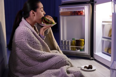 Photo of Young woman eating burger near refrigerator in kitchen at night. Bad habit