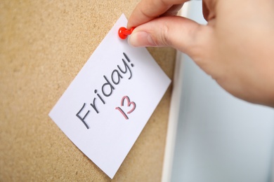 Photo of Woman pinning paper note with phrase Friday! 13 to cork board, closeup. Bad luck superstition