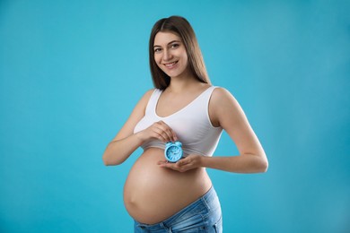 Young pregnant woman holding alarm clock near her belly on light blue background. Time to give birth