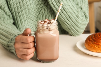 Woman holding mason jar of delicious cocoa drink with marshmallows at white table, closeup