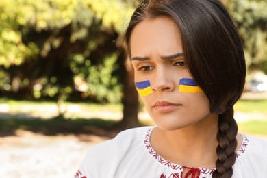Photo of Sad young woman with drawings of Ukrainian flag on face in park, space for text
