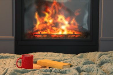 Cup of hot drink and books on knitted blanket near fireplace at home, space for text. Cozy atmosphere