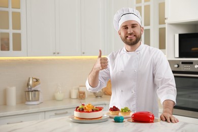 Photo of Happy professional confectioner with delicious cakes showing thumb up gesture in kitchen