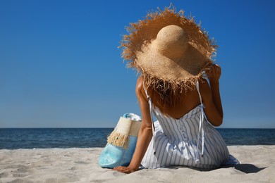 Woman with beach bag and straw hat lying on sand near sea, back view