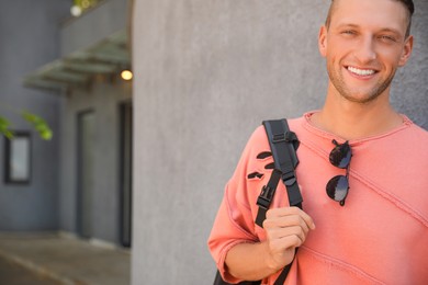Handsome young man with stylish sunglasses and backpack near grey wall outdoors, space for text