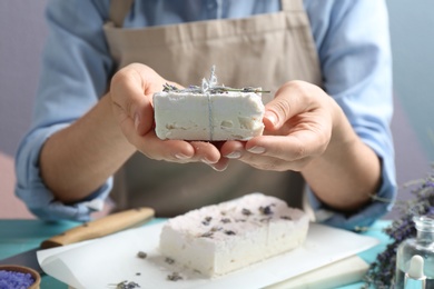 Woman holding hand made soap bar with lavender flowers at light blue wooden table, closeup
