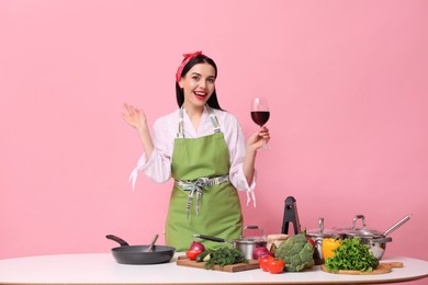 Young housewife with glass of wine, vegetables and different utensils on pink background