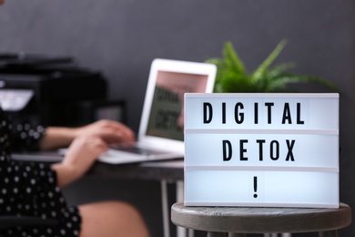 Photo of Woman using laptop at table in office, focus on lightbox with phrase DIGITAL DETOX