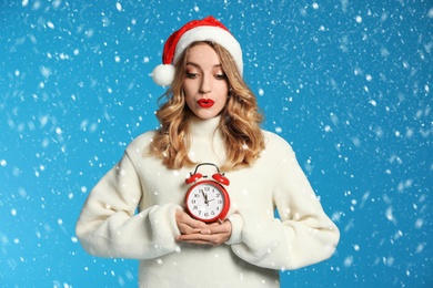 Happy young woman wearing Santa hat with alarm clock on light blue background. Christmas time