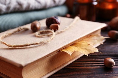 Photo of Book with autumn leaf as bookmark, scented candles and warm sweaters on wooden table, closeup