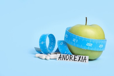 Paper with word anorexia, apple, measuring tape and pills on light blue background
