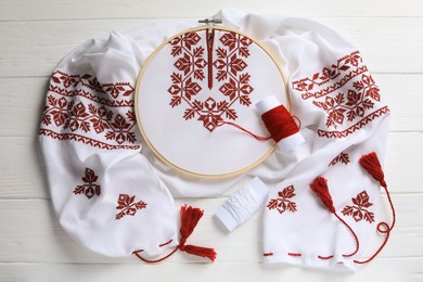 Photo of Shirt with red embroidery design in hoop, needle and threads on wooden table, flat lay. National Ukrainian clothes