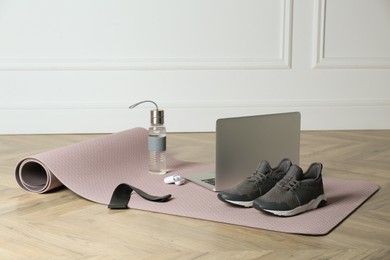 Photo of Exercise mat, laptop, bottle of water, wireless earphones, fitness elastic band and shoes on wooden floor indoors