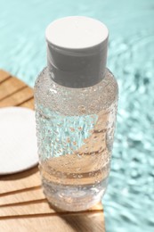 Wet bottle of micellar water and cotton pad on wooden board, closeup