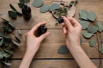 Woman taking eucalyptus essential oil with dropper from bottle at wooden table, top view
