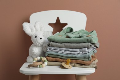 Stack of baby clothes, pacifier and toys on chair near light brown wall