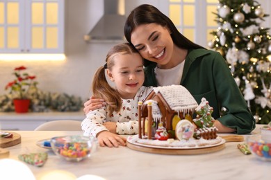 Mother and daughter with gingerbread house at table indoors