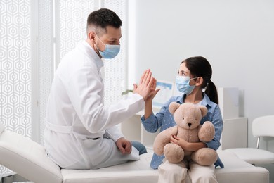 Pediatrician giving high five to little girl in hospital. Doctor and patient wearing protective masks