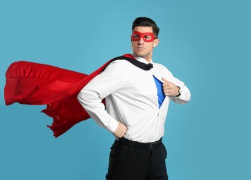Businessman in superhero cape and mask taking shirt off on light blue background