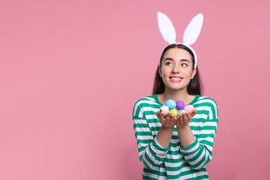 Photo of Happy woman in bunny ears headband holding painted Easter eggs on pink background. Space for text.